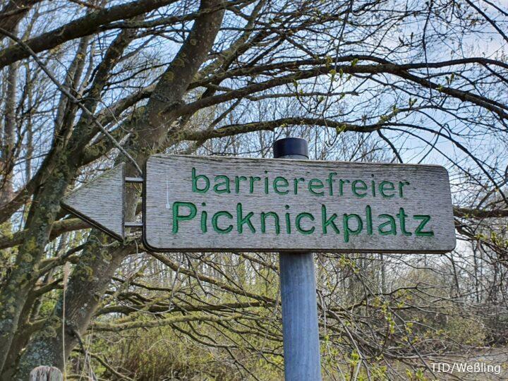 Holzschild mit grüner Aufschrift „barrierefreier Picknickplatz“ und Pfeil nach links, vor kahlen Ästen und Frühlingsknospen.