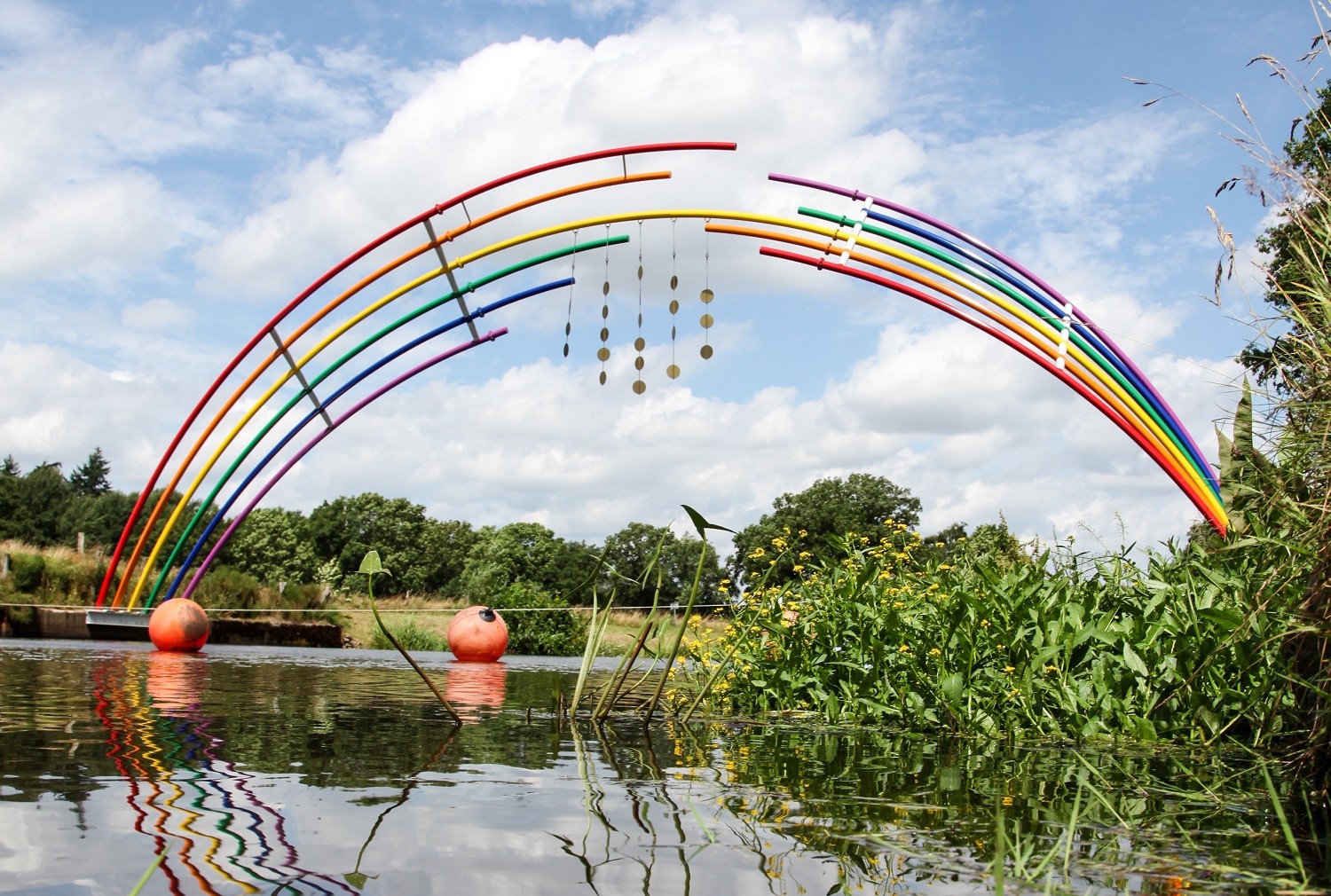 Ein Bogen aus Metallrohren in Regenbogenfarben über einen FLuss mit grünen Pflanzen an der Seite