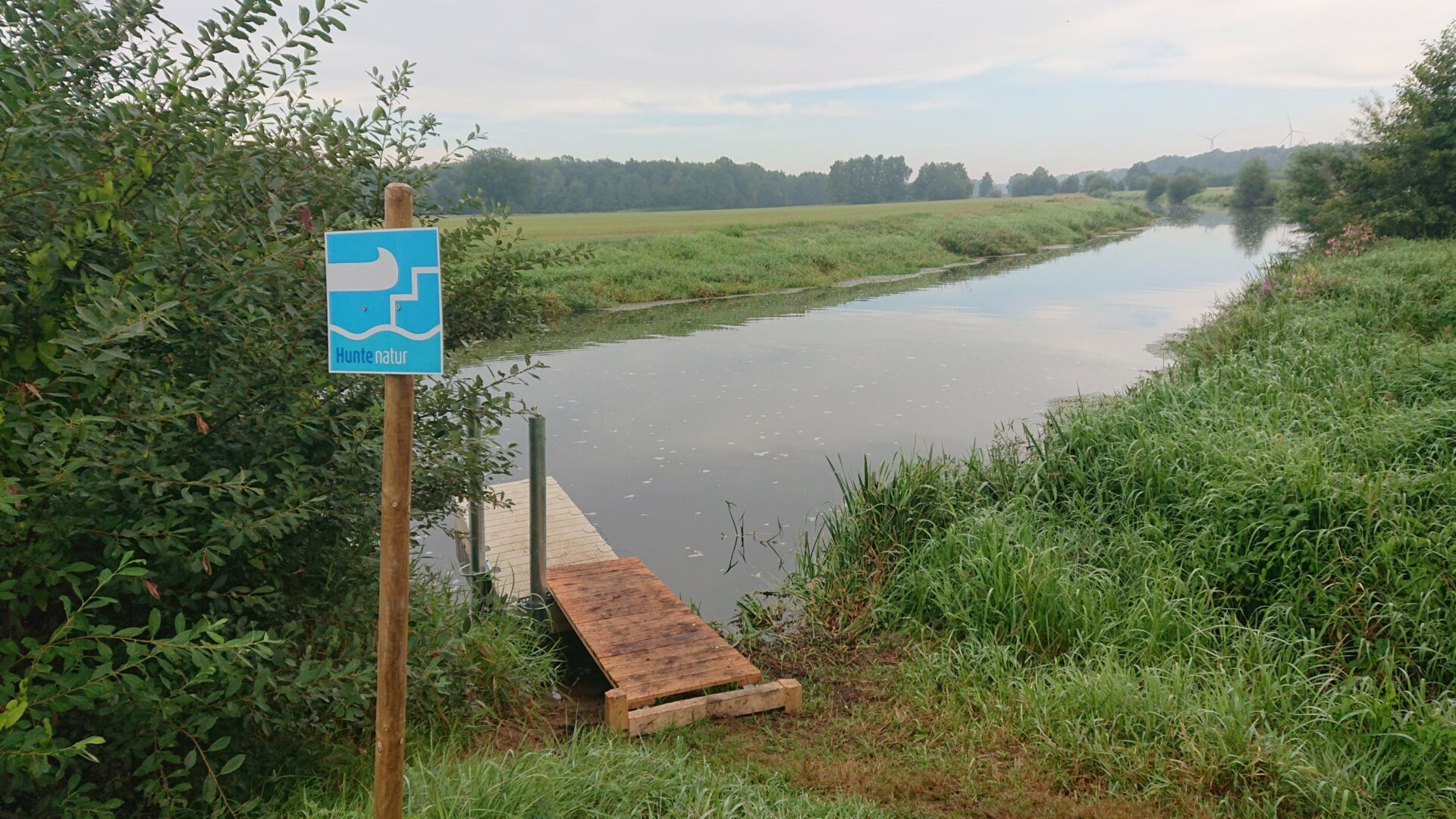 Ein kleiner Steg aus Holz an der Hunte, neben dem Steg ist ein grüner Busch und ein Pfahl mit einem blauen Schild mit einem weißen Kanu drauf