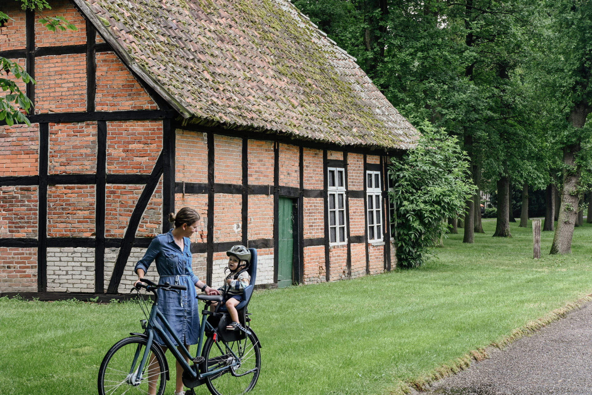 Eine Frau steht vor einem Fachwerkhaus mit einem Fahrrad, auf dem im Kindersitz ein Kleinkind sitzt.