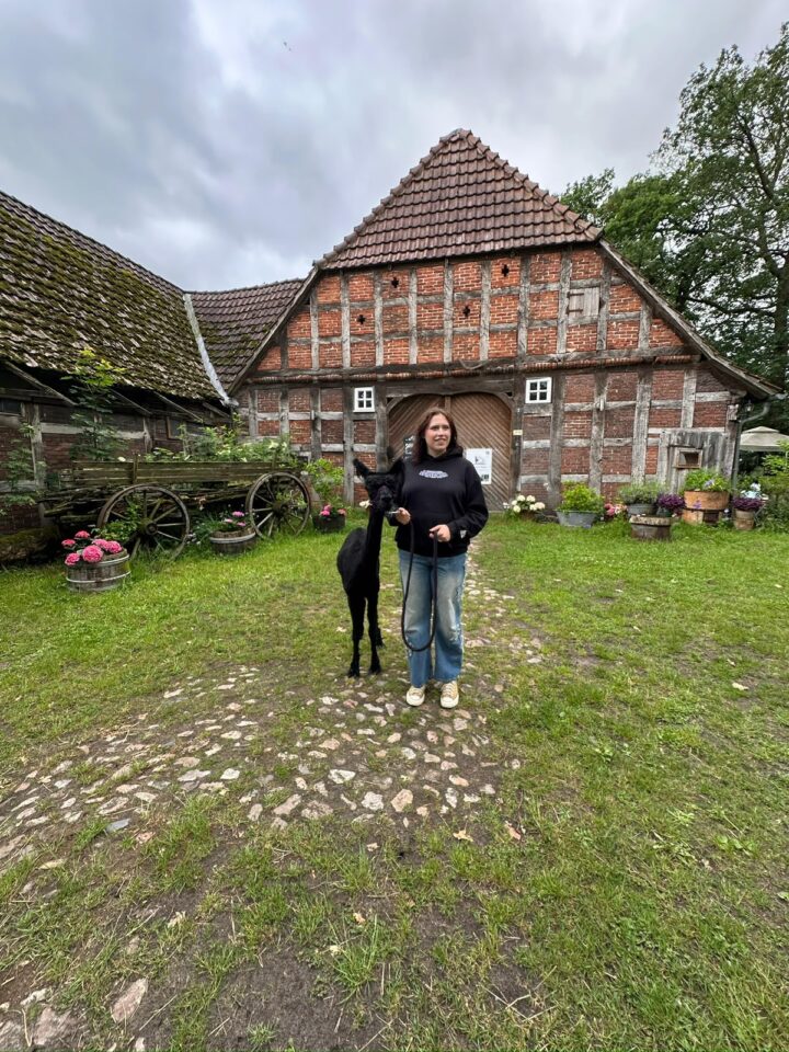 EIne junge Frau steht mit einem schwarzen Alpaka neben sich auf einem Rasen vor einem Fachwerkhaus