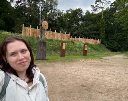 EIne junge Frau im Vordergrund schaut in die Kamera, im Hintergrund ist die Ringwallanlage der Arkeburg in Goldenstedt zu sehen.