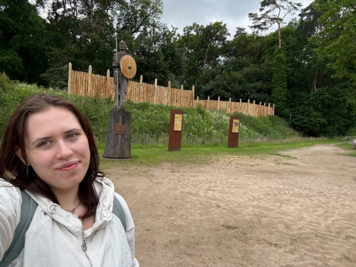 EIne junge Frau im Vordergrund schaut in die Kamera, im Hintergrund ist die Ringwallanlage der Arkeburg in Goldenstedt zu sehen.