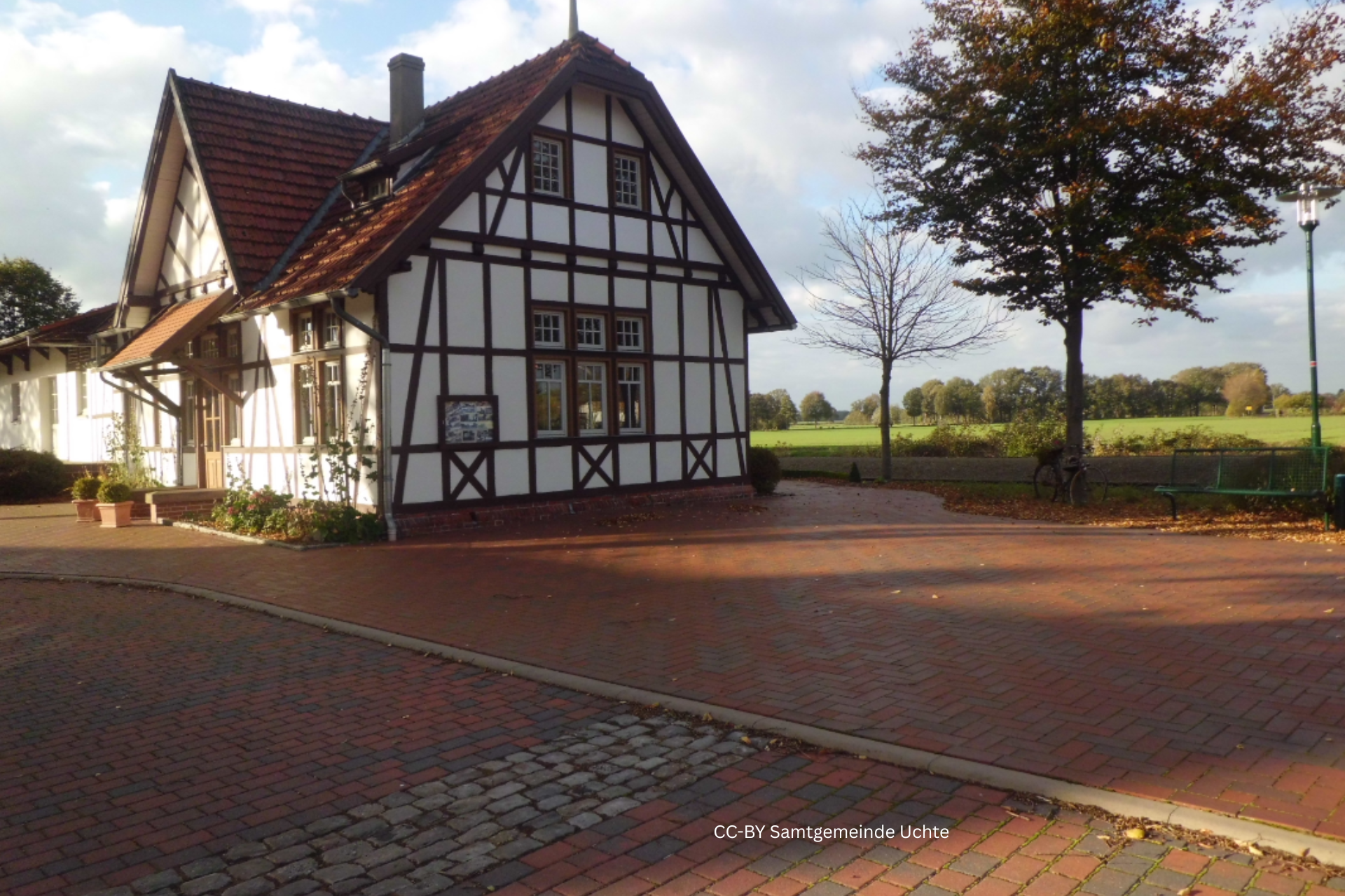 Fachwerkgebäude des ehemaligen Bahnhofs Lavelsloh mit rotem Ziegeldach, umgeben von gepflastertem Vorplatz und Blick auf angrenzende Wiesenlandschaft.