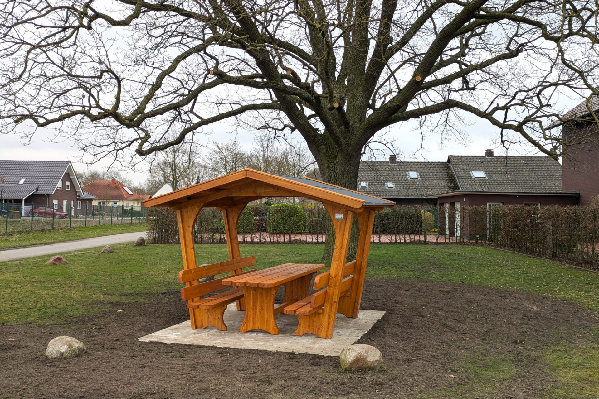MarlerEiche Ein überdachter Picknickplatz aus Holz mit zwei Bänken und einem Tisch auf einem grasbewachsenen Grundstück mit einer großen Eiche im Hintergrund