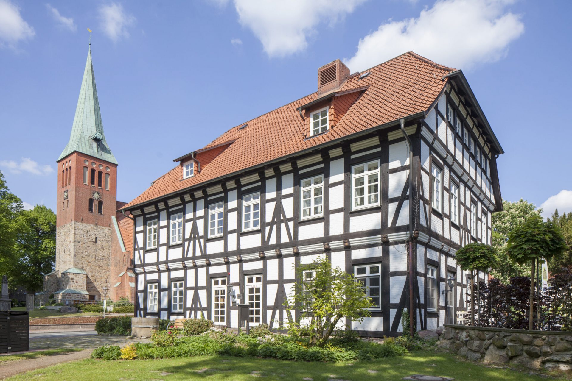 Ein großes weißes Fachwerkhaus mit roten Dach mit grüner Wiese davor und einem Kirchturm im Hintergrund